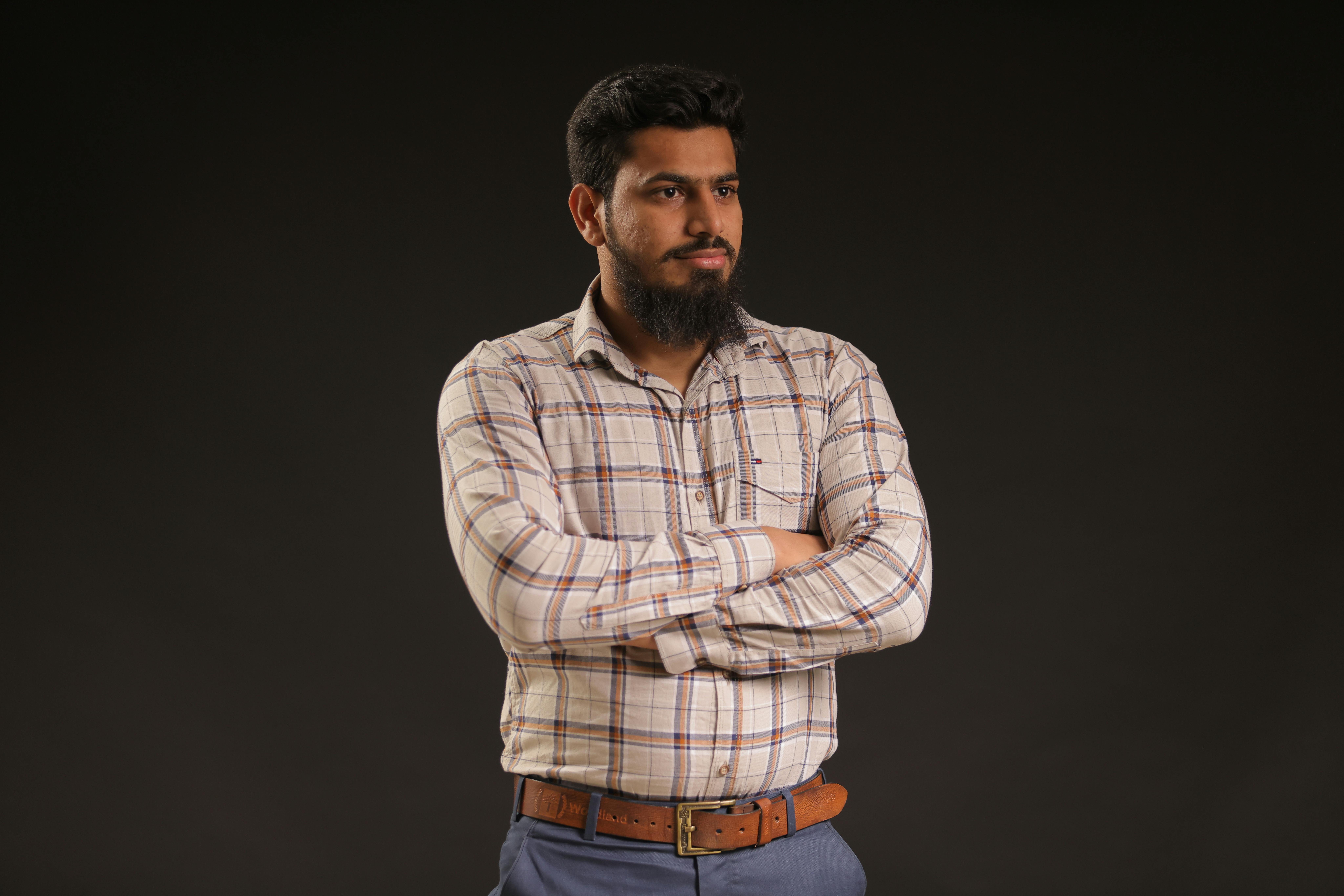 Man with a beard and moustache confidently standing with arms crossed in a studio setting.